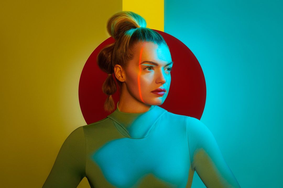 Portrait of a woman with fair skin and blonde hair against colorful yellow and turquoise backdrop with red circle accent in the background.